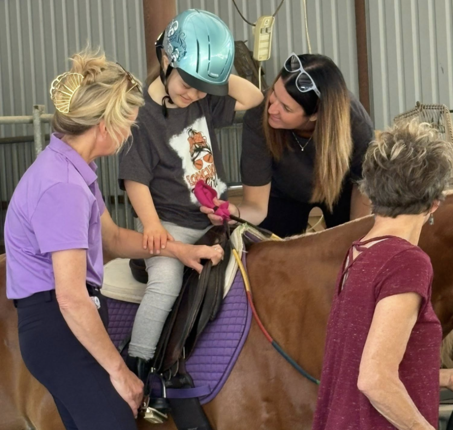 A smiling young rider on a chestnut horse, guided by instructors